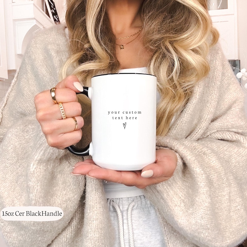 Puede incluir: Taza de cer&aacute;mica blanca con asa y borde negros, con el texto "your custom text here" y un s&iacute;mbolo de coraz&oacute;n. La taza la sostiene una persona que lleva anillos dorados y un su&eacute;ter beige. La taza est&aacute; etiquetada como "15oz Cer BlackHandle".