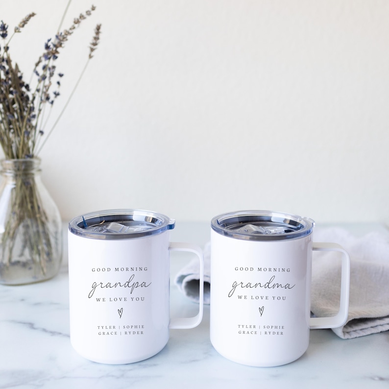 Puede incluir: Dos tazas blancas aisladas con tapas y asas negras. Las tazas tienen el texto "GOOD MORNING grandpa WE LOVE YOU" y "GOOD MORNING grandma WE LOVE YOU" impreso, junto con nombres. Un jarr&oacute;n de lavanda est&aacute; en el fondo.