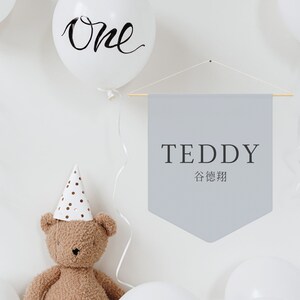 May include: A gray banner with the text "TEDDY" and the Chinese characters for "Teddy Bear" hanging on a white wall. There is a white balloon with the word "One" written on it, a brown teddy bear wearing a white and gold polka dot party hat, and white balloons in the background.