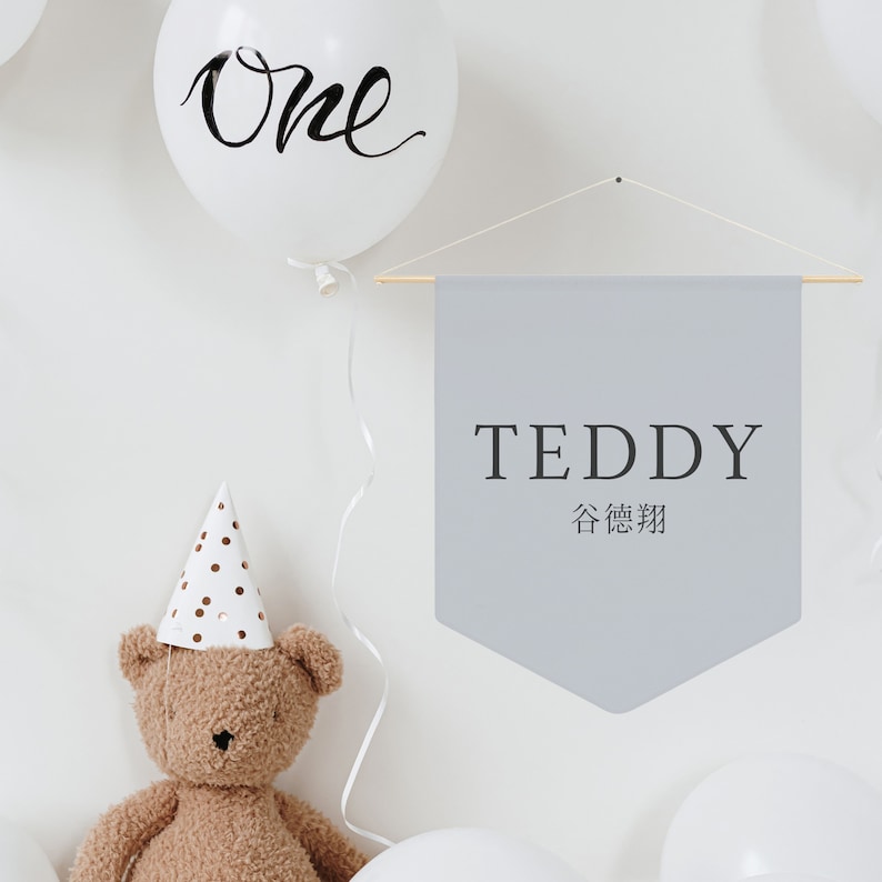 May include: A gray banner with the text "TEDDY" and the Chinese characters for "Teddy Bear" hanging on a white wall. There is a white balloon with the word "One" written on it, a brown teddy bear wearing a white and gold polka dot party hat, and white balloons in the background.