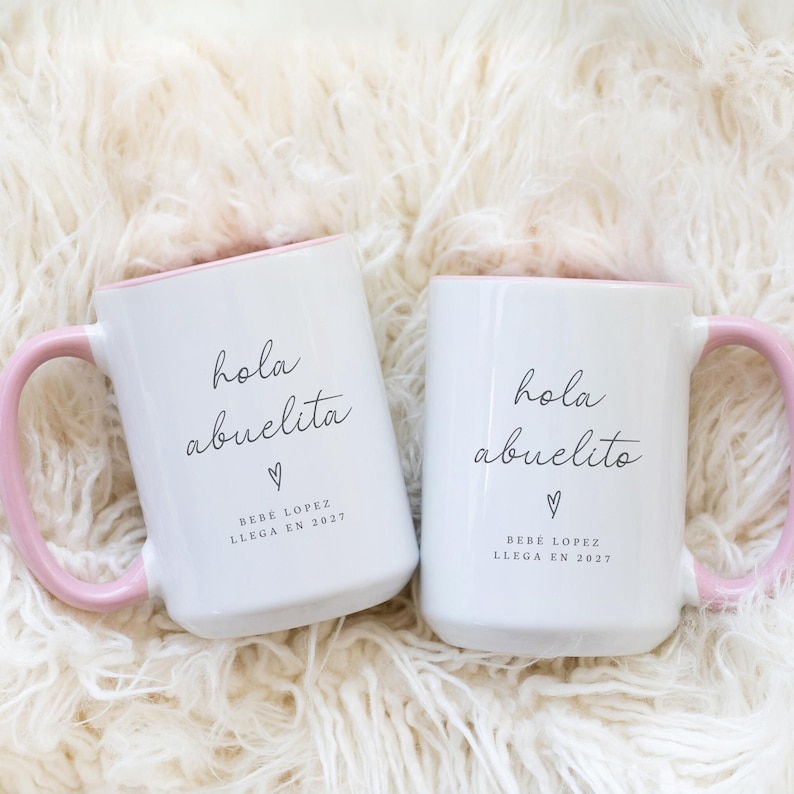 May include: Two white ceramic mugs with pink handles and rims. The mugs have the text "hola abuelita" and "hola abuelito" in black script, with a heart symbol and the text "BEBE LOPEZ LLEGA EN 2027".