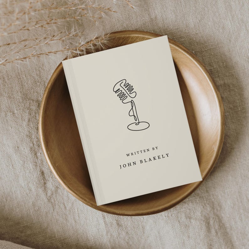 May include: A cream-colored book with a minimalist black line drawing of a microphone on the cover. The text "WRITTEN BY JOHN BLAKELY" is below the microphone. The book rests in a wooden bowl.