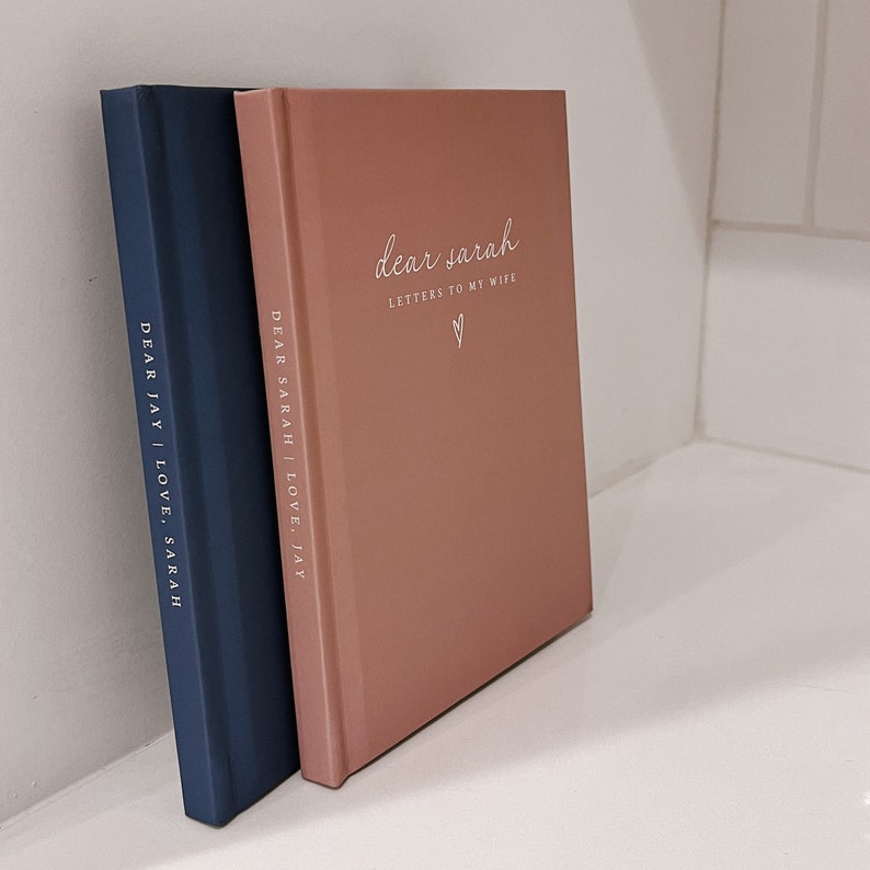 May include: Two hardcover journals, one navy blue and one dusty rose, lean against a white surface. The dusty rose journal reads "dear sarah Letters to my wife" with a heart symbol. The navy journal reads "DEAR JAY | LOVE, SARAH".
