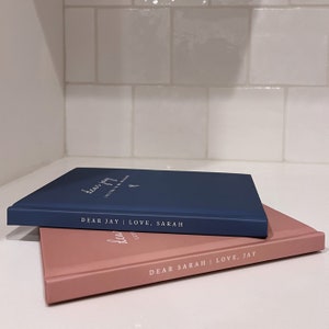May include: Two hardcover journals, one navy blue and one dusty rose, are stacked. The navy journal reads "Dear Jay | Love, Sarah" in white script. The dusty rose journal reads "Dear Sarah | Love, Jay".