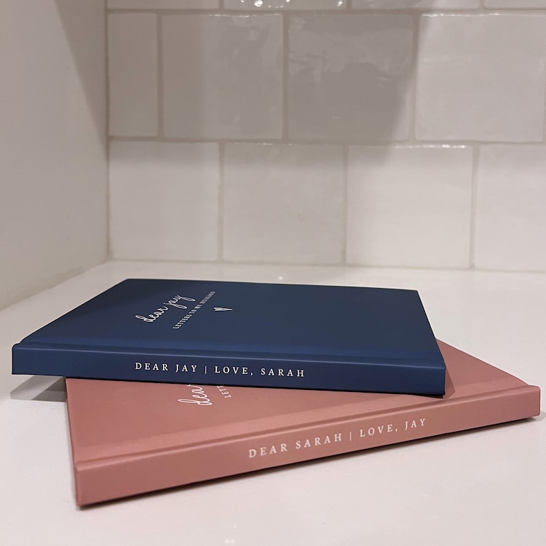 May include: Two hardcover journals, one navy blue and one dusty rose, are stacked. The navy journal reads "Dear Jay | Love, Sarah" in white script. The dusty rose journal reads "Dear Sarah | Love, Jay".