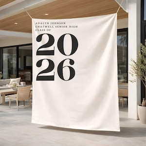 May include: Cream graduation banner with black text. The banner reads "ADALYN JOHNSON CHATWELL SENIOR HIGH CLASS OF 2026". The banner is hanging outdoors, with a patio and furniture visible in the background.