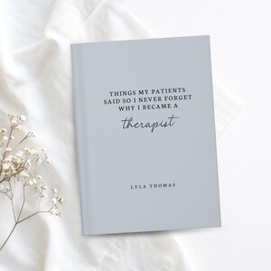 May include: A light blue journal with the text "THINGS MY PATIENTS SAID SO I NEVER FORGET WHY I BECAME A therapist" and the author's name, Lyla Thomas. The journal is on a white surface with a sprig of white flowers.