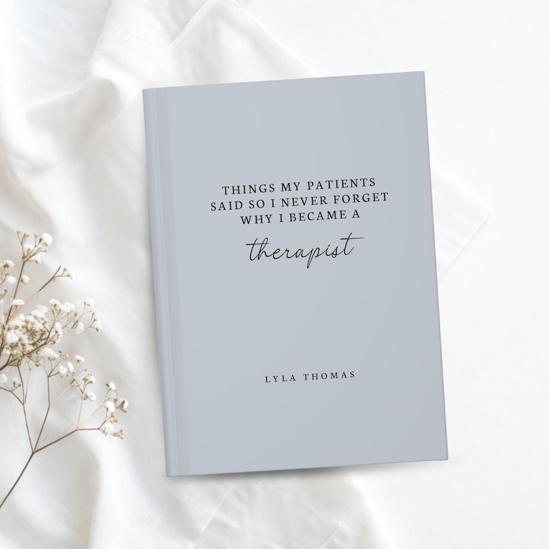 May include: A light blue journal with the text "THINGS MY PATIENTS SAID SO I NEVER FORGET WHY I BECAME A therapist" and the author's name, Lyla Thomas. The journal is on a white surface with a sprig of white flowers.