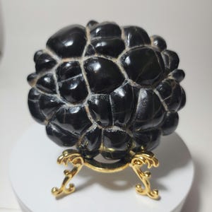 Op de afbeelding: Een zwarte obsidiaans bol met een getextureerd oppervlak, dat lijkt op een schildpadden schild, getoond op een gouden standaard.