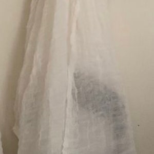 May include: White scarf with a patterned border in shades of brown, tan, and blue. The scarf is hanging on a black hanger against a white wall. The scarf appears to be made of a lightweight fabric.