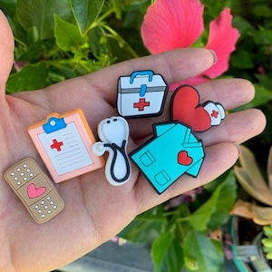 May include: A set of six colorful medical-themed shoe charms. The charms include a red heart, a first aid kit, a stethoscope, a bandage, a doctor's coat, and a clipboard with a red cross.