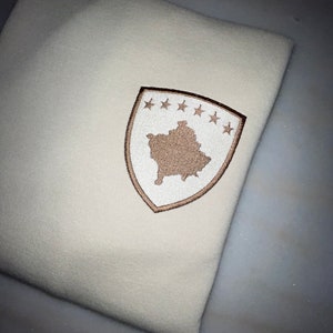 Może przedstawiać: Biały sweter z brązowym haftowanym herbem przedstawiającym zarys Kosowa i sześć gwiazd.