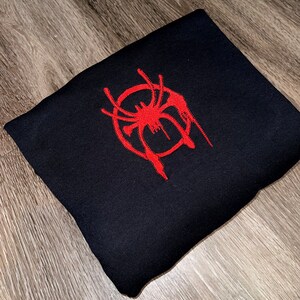 Puede incluir: Sudadera negra con un logotipo de Spider-Man bordado en rojo. El logotipo es una telara&ntilde;a estilizada con un efecto de goteo.