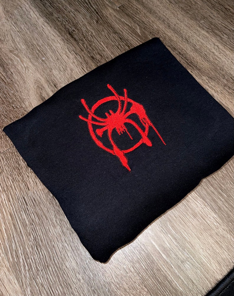 Puede incluir: Sudadera negra con un logotipo de Spider-Man bordado en rojo. El logotipo es una telara&ntilde;a estilizada con un efecto de goteo.
