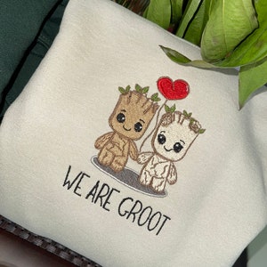 May include: A beige sweatshirt with an embroidered design of two cartoon Groot characters holding hands. One Groot is holding a red heart balloon. The text "We are Groot" is embroidered below the characters.