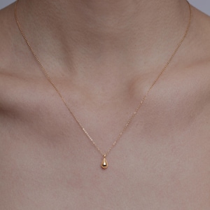 Può includere: Una delicata collana d'oro con un piccolo pendente a forma di goccia. Il pendente è sospeso a una catena sottile, creando un look minimalista ed elegante. La collana è mostrata su uno sfondo color carne.