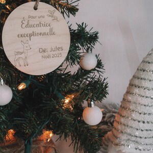 Peut inclure: Ornement de Noël en bois avec le texte "Pour une Educatrice exceptionnelle De: Jules Noël 2025" et une illustration de cerf. L'ornement est suspendu à un sapin décoré de boules blanches et de lumières.