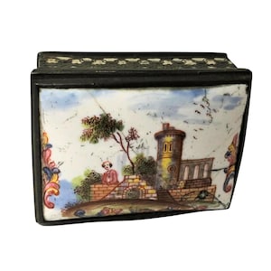 May include: A rectangular box with a dark frame and a white enamel lid featuring a hand-painted landscape scene. The scene depicts a stone tower, a brick wall, a person, and trees under a blue sky. The lid has decorative floral accents on the sides.