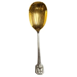 May include: A silver-toned serving spoon featuring a large, gold-washed bowl with a fluted, shell-like design. The handle is slender and vertical-ribbed, terminating in a rectangular base decorated with a relief pattern of three small fruits.