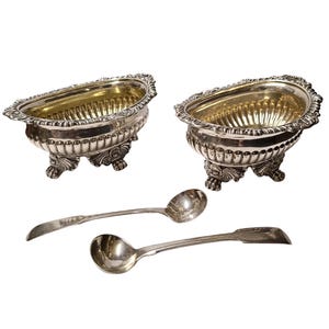 May include: Two silver-toned oval bowls with fluted sides and ornate scalloped rims, each supported by three decorative lion-paw feet. Two matching silver-toned spoons with rounded bowls and flat, tapered handles accompany the two bowls.