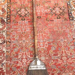 May include: A long-handled push broom featuring a weathered wooden handle, a metallic head attachment secured with rivets, and stiff, dark brown natural bristles held together by a single horizontal wire wrap near the base of the brush head.