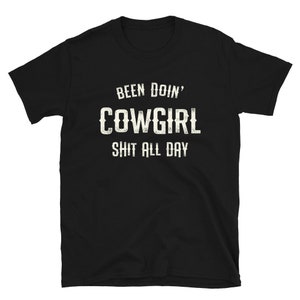 May include: Black t-shirt with white text that reads "Been Doin' Cowgirl Shit All Day".
