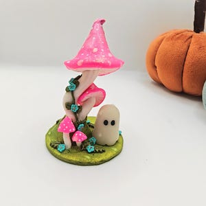 May include: A small figurine featuring a tall, bright pink mushroom with white spots, accompanied by two smaller pink mushrooms and a tiny, smooth, off-white ghost character with two black eyes, all set on a textured green base with blue flowers.