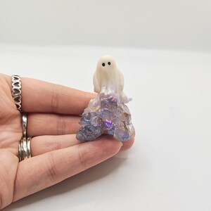 May include: A small, white ghost figurine with black dot eyes and a blush of pink on its cheeks. The ghost sits atop a cluster of iridescent, multi-colored crystals. The figurine is held in a hand against a white background.