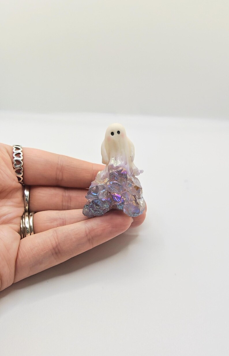 May include: A small, white ghost figurine with black dot eyes and a blush of pink on its cheeks. The ghost sits atop a cluster of iridescent, multi-colored crystals. The figurine is held in a hand against a white background.
