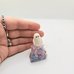 May include: A small, white ghost figurine with black dot eyes and a blush sits atop a colorful, iridescent crystal formation. The crystal has shades of purple, blue, and gold. The figurine is a whimsical, decorative item.