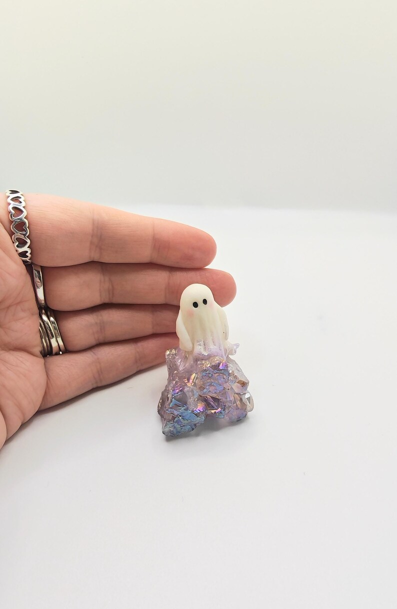 May include: A small, white ghost figurine with black dot eyes and a blush sits atop a colorful, iridescent crystal formation. The crystal has shades of purple, blue, and gold. The figurine is a whimsical, decorative item.