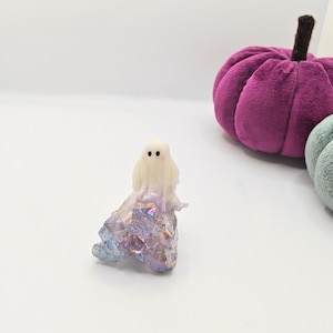 May include: A small, white ghost figurine with black dot eyes and a light purple and blue crystal base. A plush, purple pumpkin with a brown stem is in the background. The figurine is a Halloween decoration.