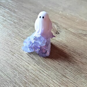 May include: A whimsical ghost figurine with a white body and black dot eyes. The ghost's base is made of sparkling purple crystals. The figurine is set on a wooden surface, creating a charming, spooky aesthetic.