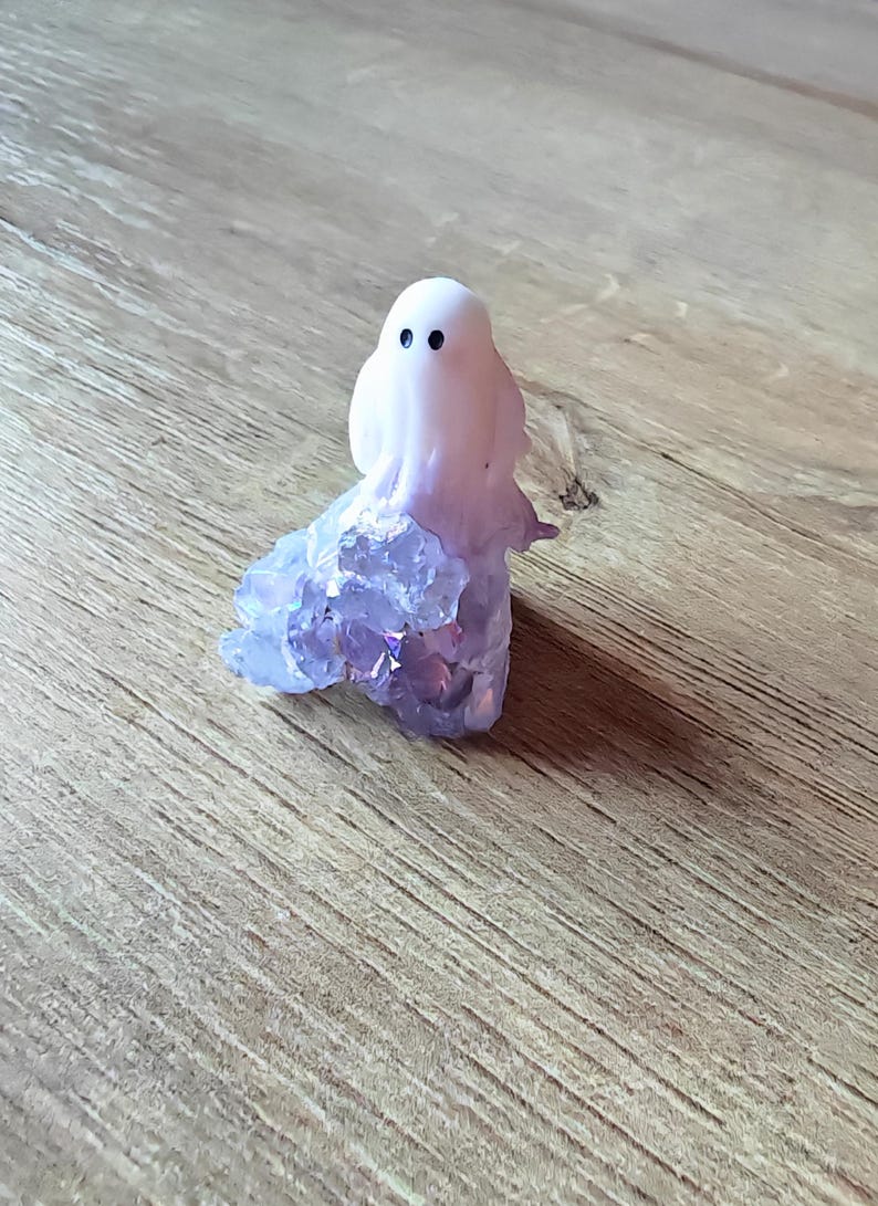 May include: A whimsical ghost figurine with a white body and black dot eyes. The ghost's base is made of sparkling purple crystals. The figurine is set on a wooden surface, creating a charming, spooky aesthetic.