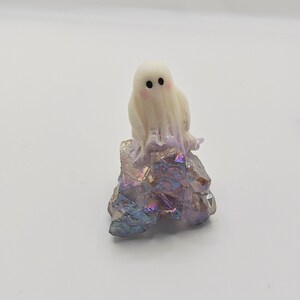 May include: A small, white ghost figurine with black eyes and rosy cheeks sits atop a cluster of iridescent crystals. The crystals display shades of purple, blue, and gold. This decorative item is perfect for Halloween or as a unique home accent.