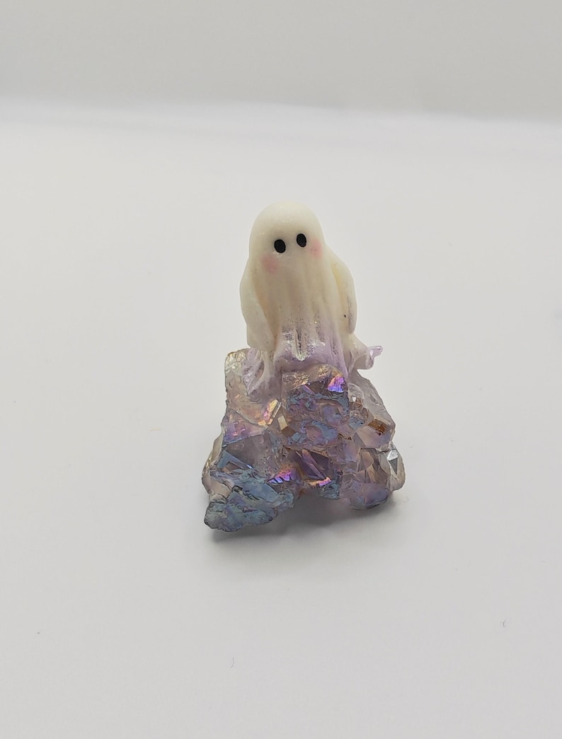 May include: A small, white ghost figurine with black eyes and rosy cheeks sits atop a cluster of iridescent crystals. The crystals display shades of purple, blue, and gold. This decorative item is perfect for Halloween or as a unique home accent.