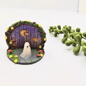 May include: A miniature fairy door with a ghost figurine. The door is framed by a brown arch with yellow flowers and green vines. The door is purple with a crescent moon and stars. Red and white mushroom accents.