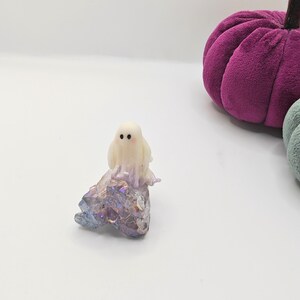 May include: A small, off-white ghost figurine with two black dot eyes sits atop a sparkling, iridescent crystal formation. A plush, purple pumpkin is in the background. The items are set against a white backdrop, evoking a Halloween theme.