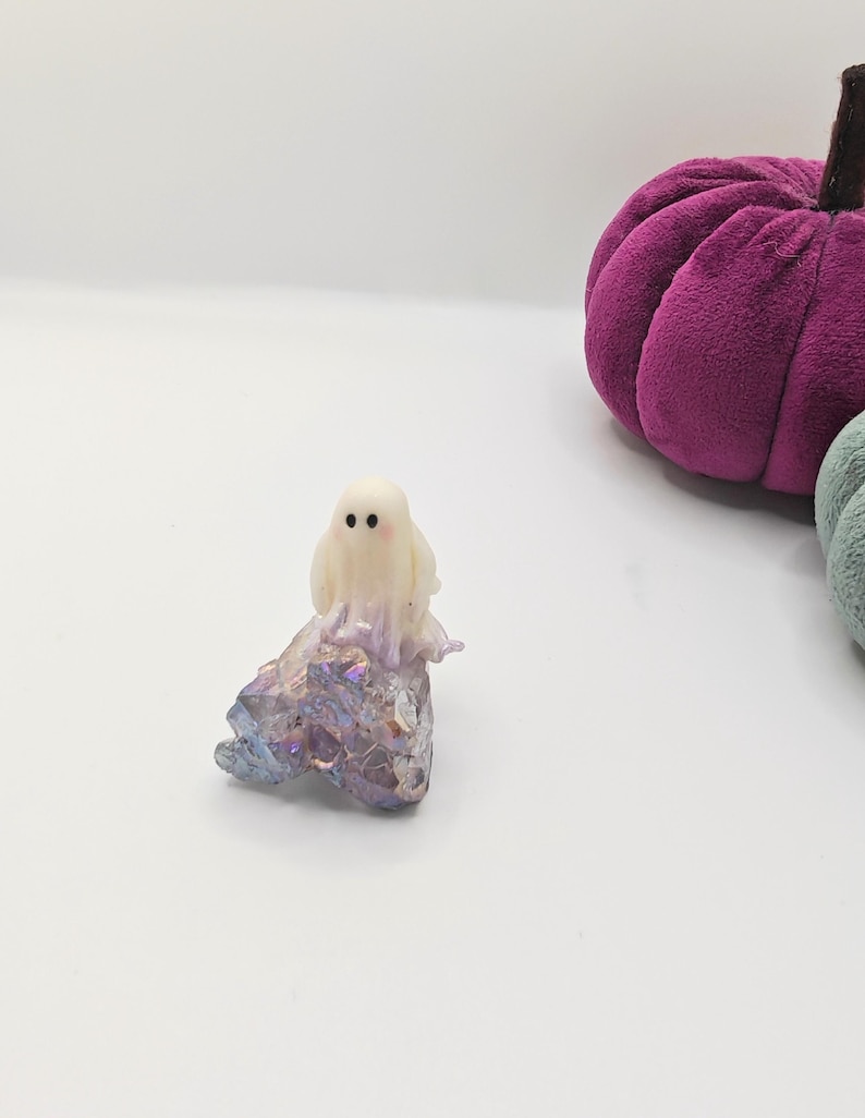 May include: A small, off-white ghost figurine with two black dot eyes sits atop a sparkling, iridescent crystal formation. A plush, purple pumpkin is in the background. The items are set against a white backdrop, evoking a Halloween theme.
