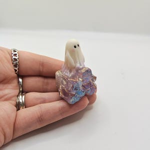 May include: A small, white ghost figurine with black dot eyes sits atop a colorful, iridescent crystal formation. The crystal base features shades of purple, pink, and blue. The ghost is a cute, whimsical design.