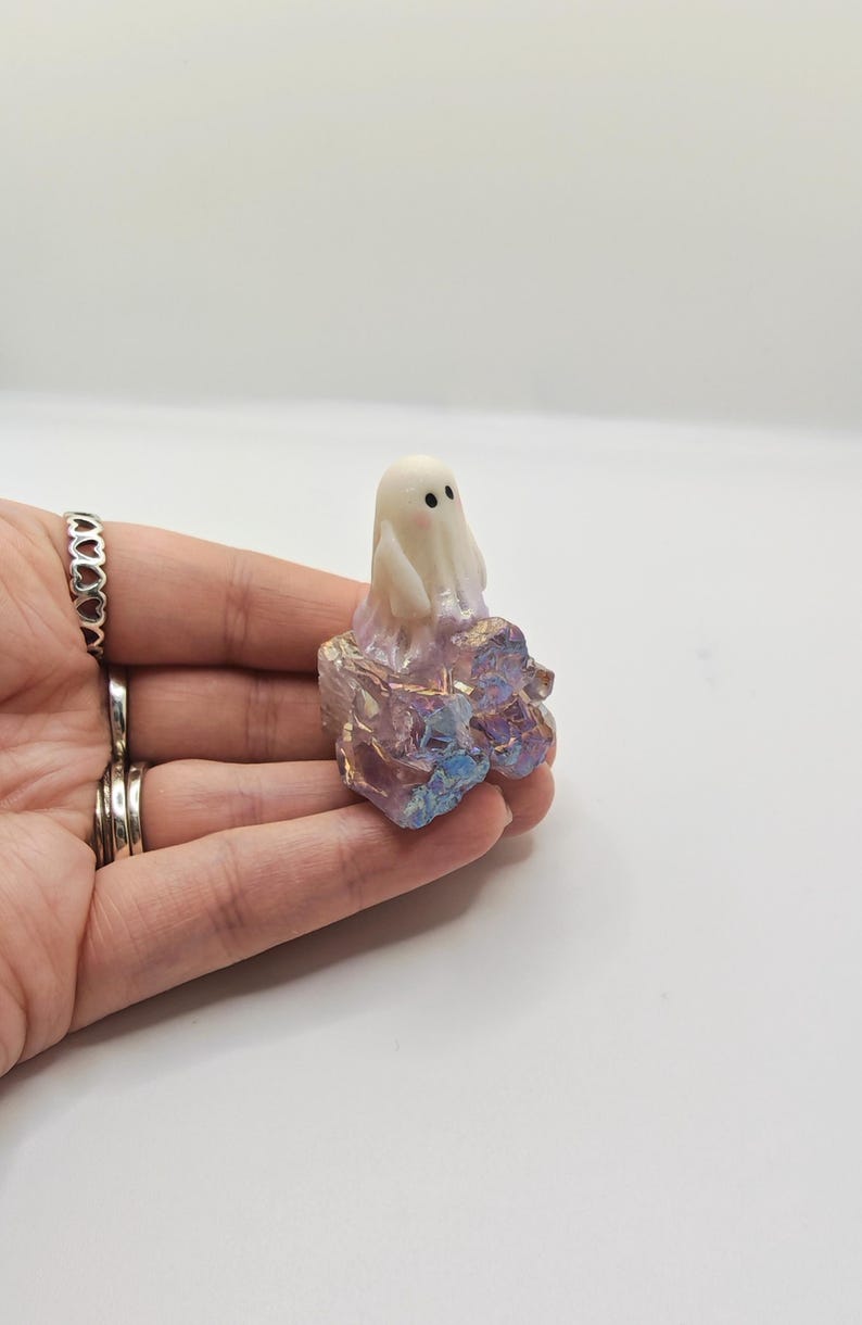 May include: A small, white ghost figurine with black dot eyes sits atop a colorful, iridescent crystal formation. The crystal base features shades of purple, pink, and blue. The ghost is a cute, whimsical design.