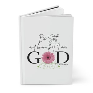 以下が含まれることがあります： ピンクの花と「Be Still and know that I am GOD」というテキスト、聖書の参照「Psalm 46:10」が書かれた白いジャーナル。