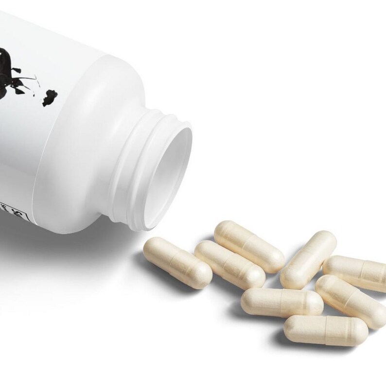 May include: A white plastic bottle with a black design on the label is open and spilling out beige capsules.
