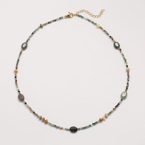 May include: A delicate necklace featuring a mix of small, colorful beads and larger oval-shaped stones in shades of green, brown, and cream. The necklace has a gold-colored chain and clasp, creating an elegant and natural look.