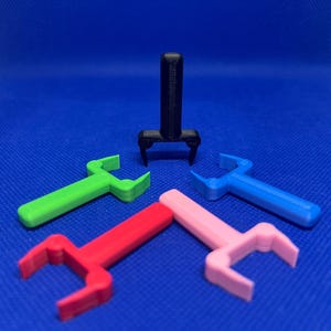 May include: Five 3D printed plastic hooks in different colors: black, green, blue, pink, and red. The hooks are designed to hang items on a wall or other surface.