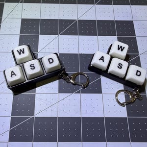 May include: Two keychains featuring the WASD keys from a computer keyboard. Each keychain has four white keys with black lettering, arranged in a cluster on a black base. The keychains are attached to silver key rings.