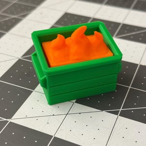 May include: A miniature green dumpster with an orange, flame-shaped object inside. The dumpster is rectangular with a hinged lid and is sitting on a black and gray checkered surface.