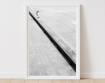 May include: Black and white framed print featuring a skateboarder on a concrete structure. The image is a high-angle shot, with the skateboarder positioned on a long, linear ledge. The print is displayed in a white frame, set against a white wall.