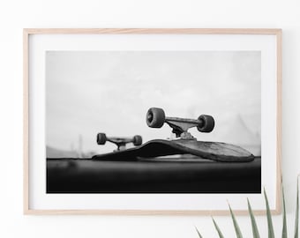 May include: Black and white framed photograph of two skateboards. The skateboards are positioned on a dark surface, with the wheels and trucks visible. The background is a blurred, overcast sky. The image is a close-up shot.