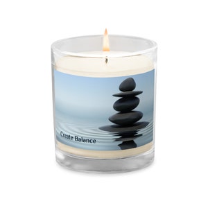 Op de afbeelding: Een glazen kaarsglas met een witte kaars en een blauwe en witte afbeelding van gestapelde stenen in water. De tekst "Create Balance" staat op het glas gedrukt.